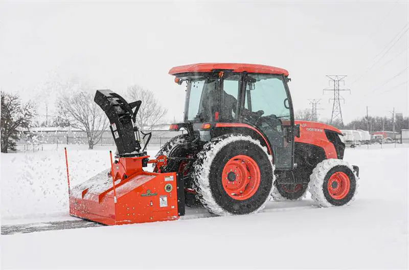 Tractor with a rear-mounted snow blower cutting through packed snow along a paved edge