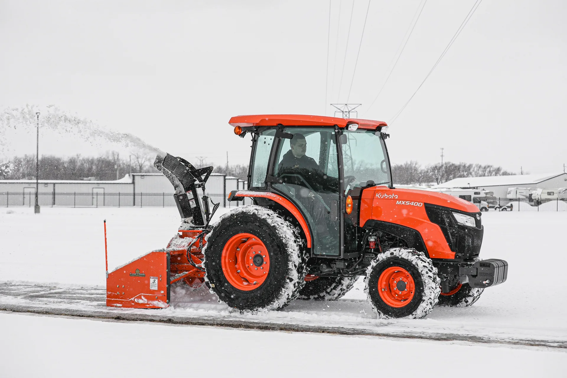 Operator in the cab of a tractor running a snow blower along a snowy path
