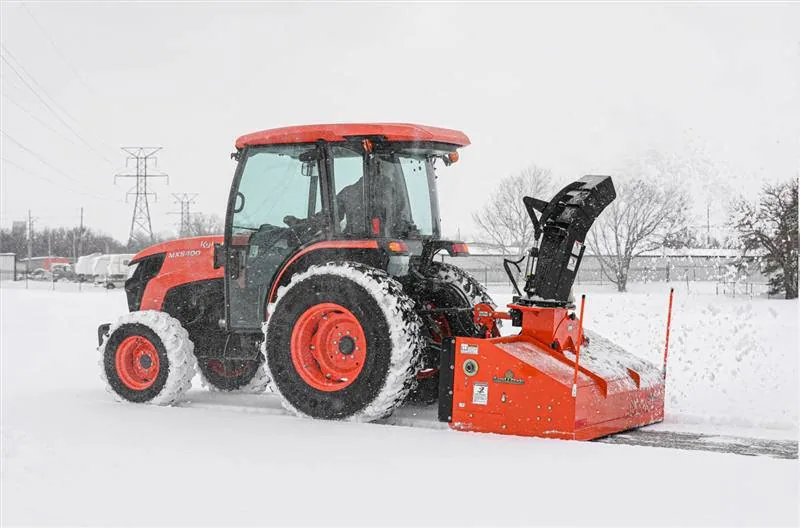 Cab tractor with a rear snow blower cutting through packed snow during a snowfall